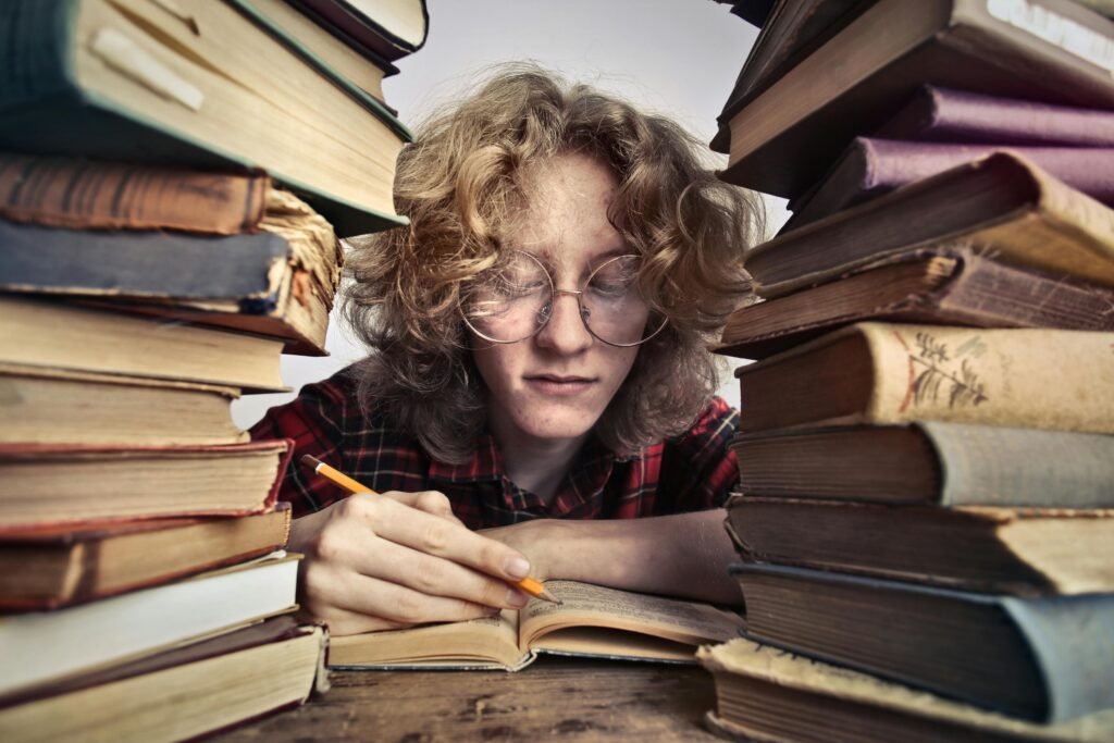 A focused young adult reading and note-taking, surrounded by stacks of books.