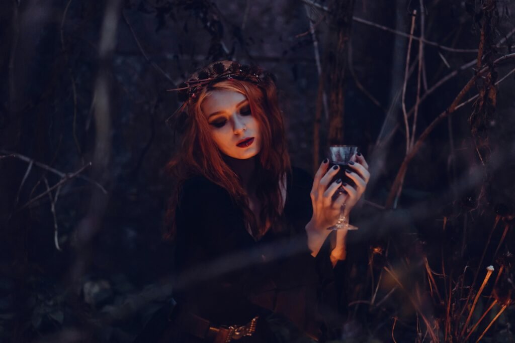A mystical woman in gothic attire holds a chalice amidst a dark, eerie forest setting.
